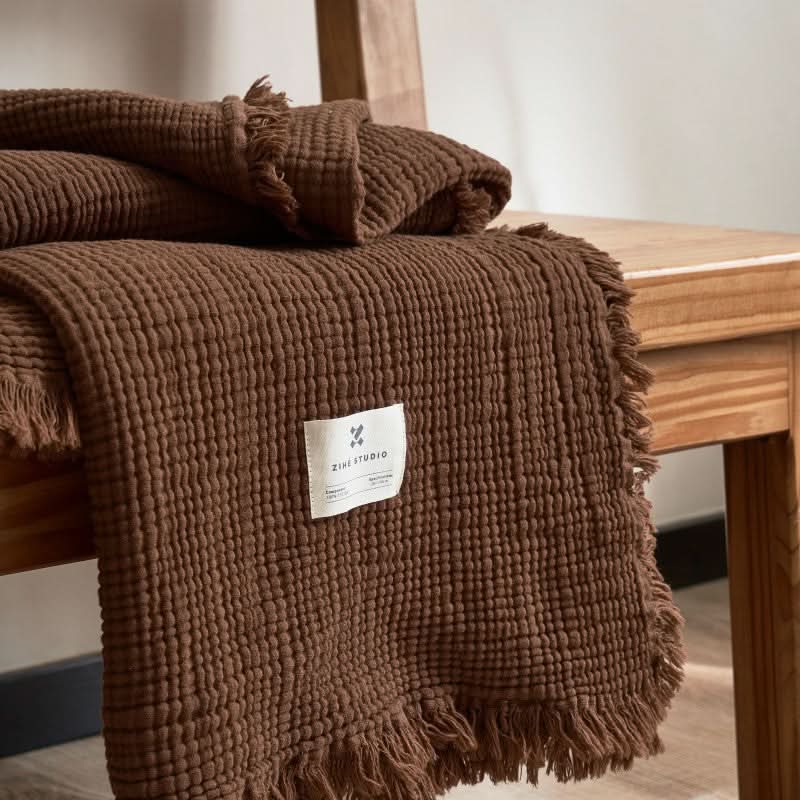 Cozy Textured Cotton Throw Blanket in rich brown, draped over a wooden chair, with tasseled edges and a brand tag.